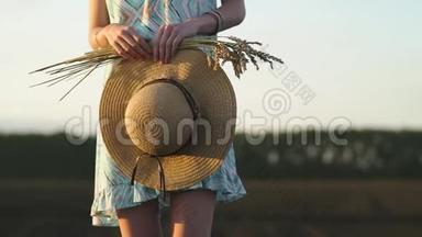 戴太阳镜的女孩站在日落时的田野里，拿着金色的麦穗和草帽。 乡村户外场景